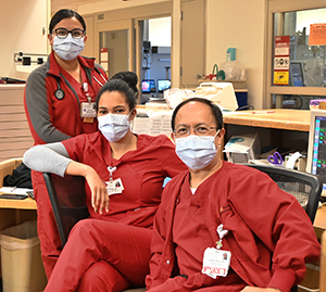 Three clinicians in protective masks sit at a nurses' station.