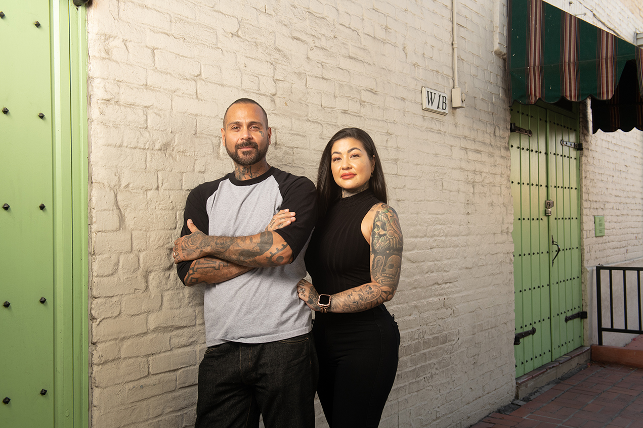 A smiling man and woman stand next to a painted brick wall