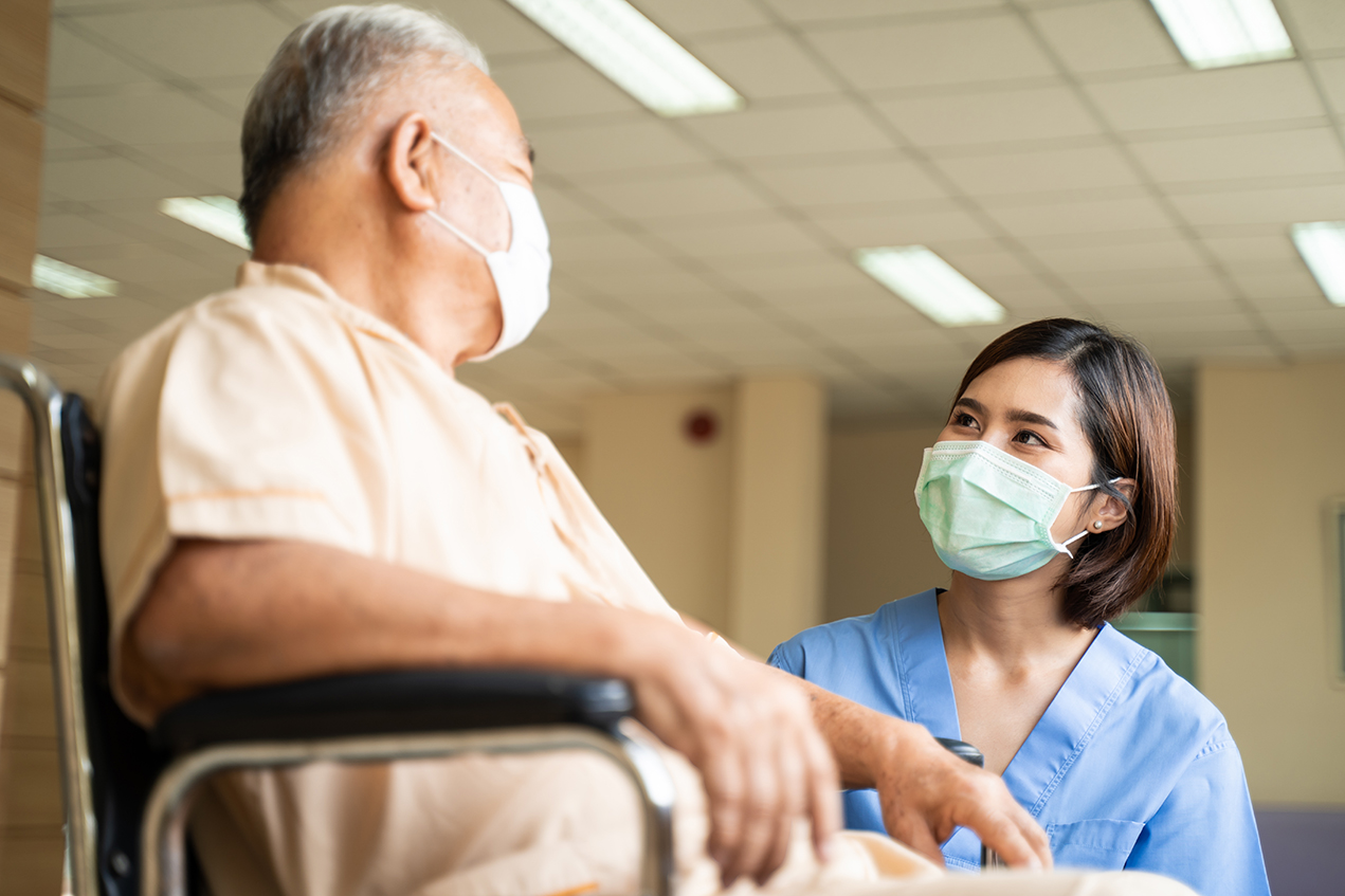 A masked older man in a wheelchair looks at a masked woman wearing scrubs