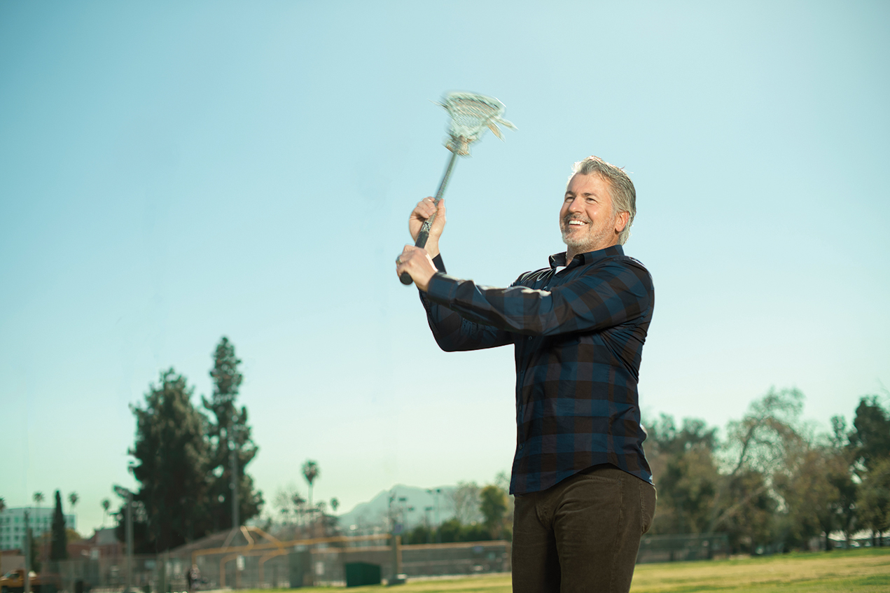A smiling man holds a lacrosse stick