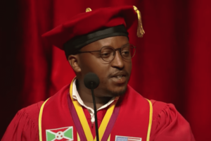A graduate in red and gold speaks at a microphone