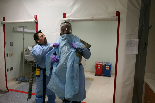 Lionel Caceres helps Robert “Bob” Vance III into personal protective equipment inside one of the rooms used for Ebola response training.  