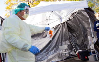 A clinician in protective gear carries a test sample past a tent.