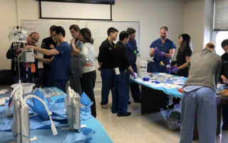 Young people in scrubs gather around tables stocked with training equipment.