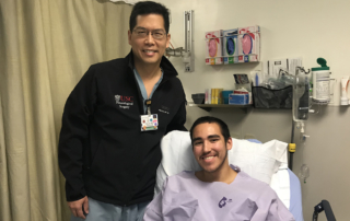A tall doctor stands next to a smiling young man sitting up in a hospital bed.