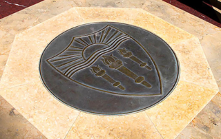 A seal in a marble floor depicts three torches burning in the light of a low sun on a shield.