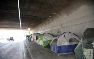 A row of tents stands sheltered by an overpass.