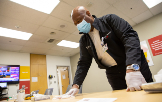 A masked and gloved man disinfects a conference room table.
