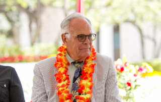 A man in his seventies sits at a ceremony wearing a lei.