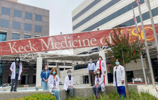Several masked health care workers stand outdoors beneath a banner reading, Keck Medicine of USC.
