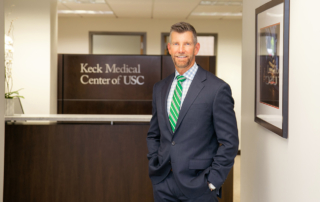 A man stands in front of a desk with a sign reading, Keck Medical Center of USC.