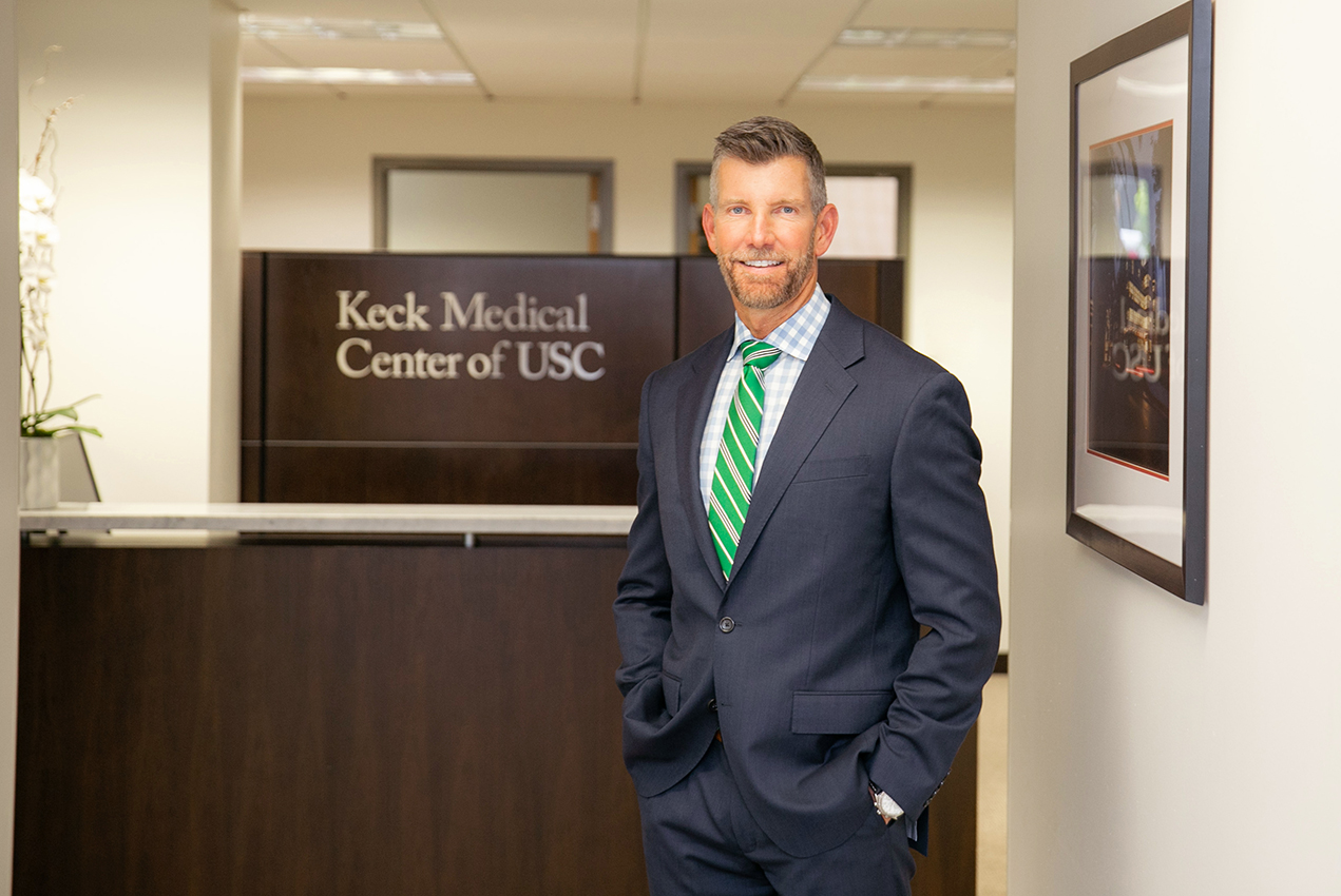 A thin man stands in front of a desk with a sign reading, Keck Medical Center of USC.