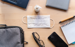 A laptop, notebook, phone, mask and hand sanitizer rest on a surface.