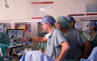Surgeons face monitors near a patient in an operating room.