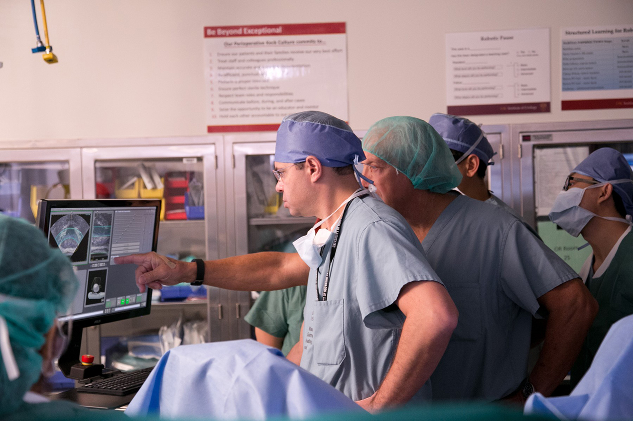 Surgeons face a monitor near a patient in an operating room.