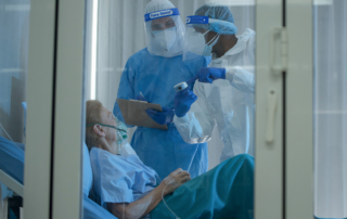 In a negative pressure room, two health care workers in full PPE stand over a patient.