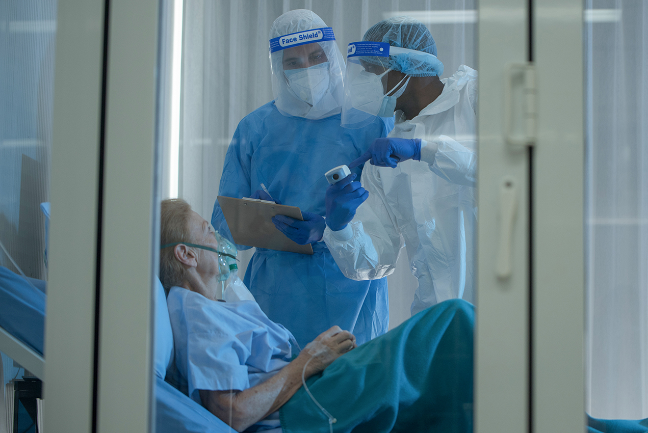 In a sealed room, two health care professionals in PPE stand over a patient in a bed.