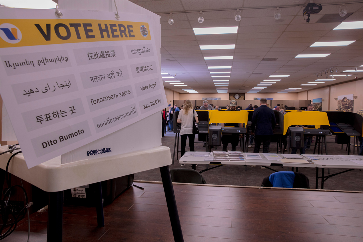A Vote Here sign in the foreground, with voters and ballot booths in the background