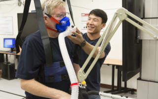 A young man assists an older man wearing a breathing apparatus.