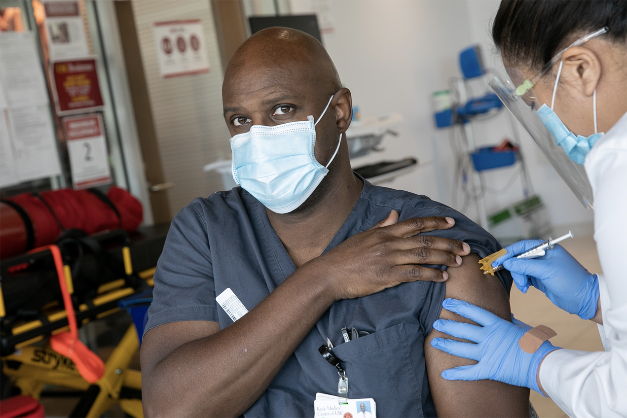 A masked physician in scrubs receives a vaccine
