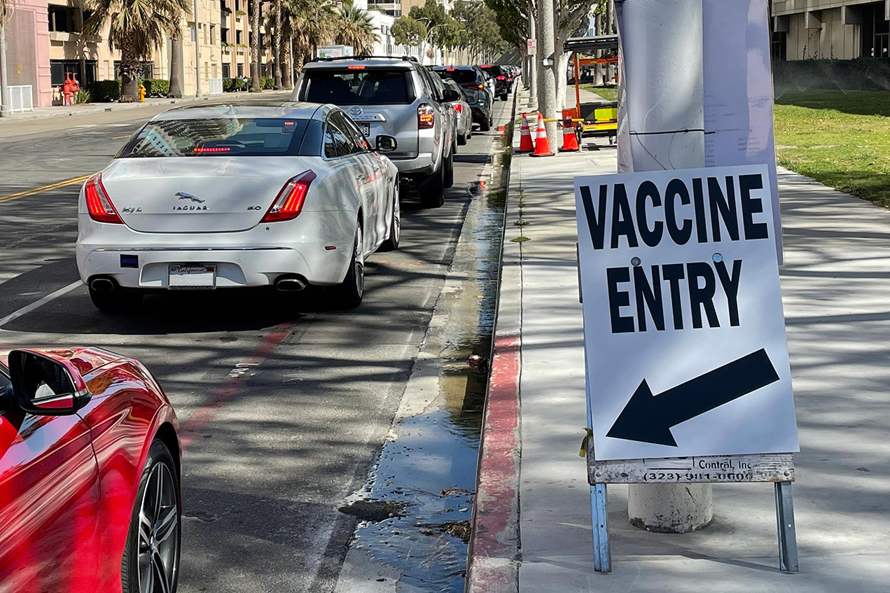 A sign advertising vaccine entry is on a sidewalk next to a line of cars