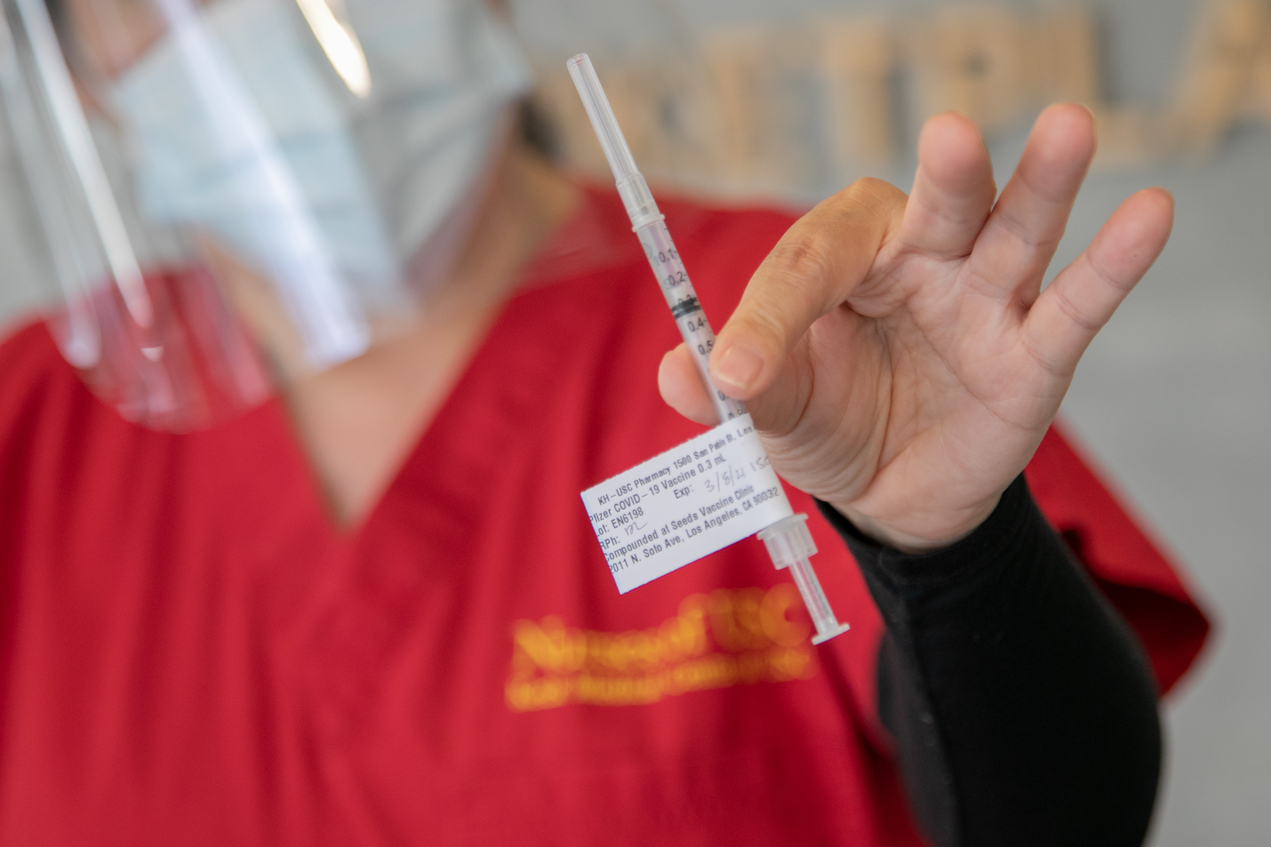 An out-of-focus person wearing red scrubs, a mask and face shield holds a syringe with a tag indicating it is a COVID-19 vaccine