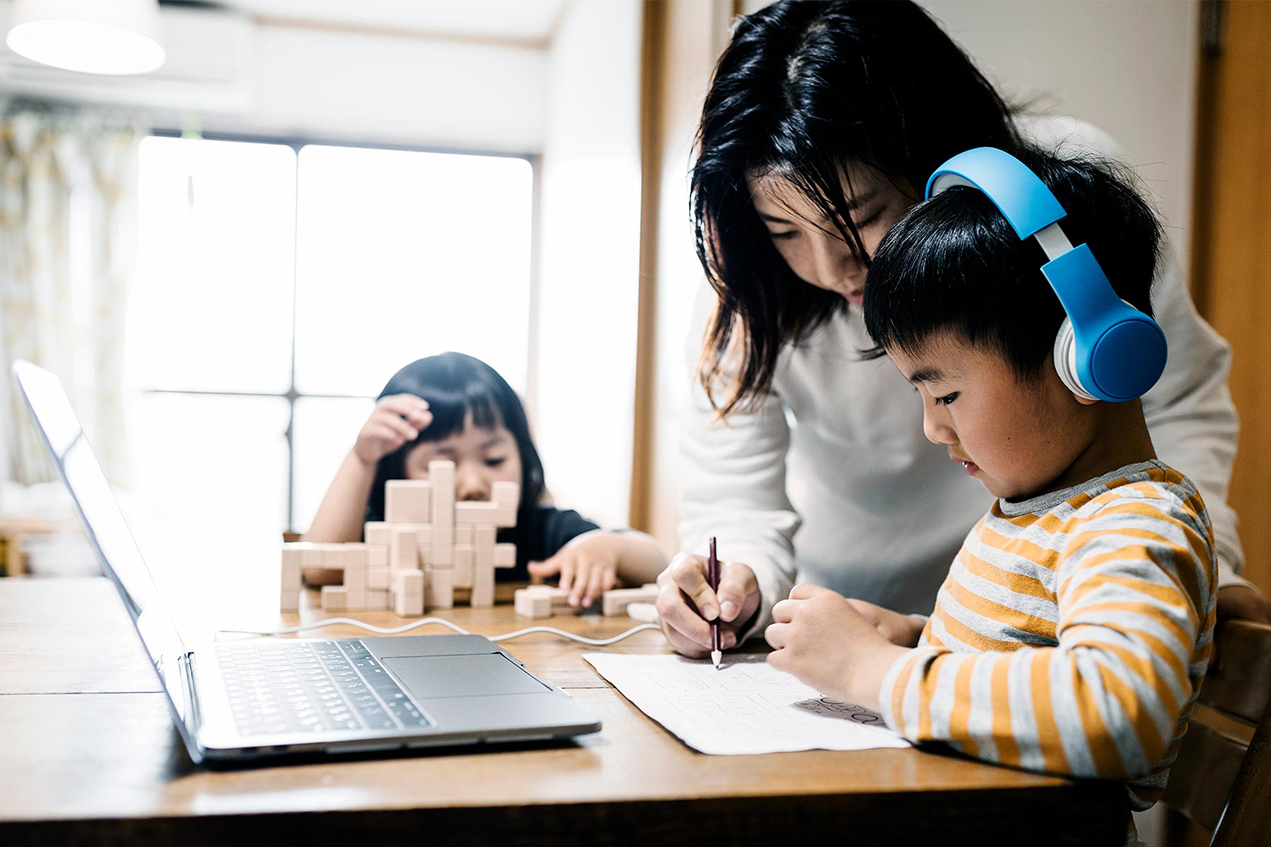 A woman leans over a child wearing headphones in front of a laptop while a younger child plays with wooden blocks