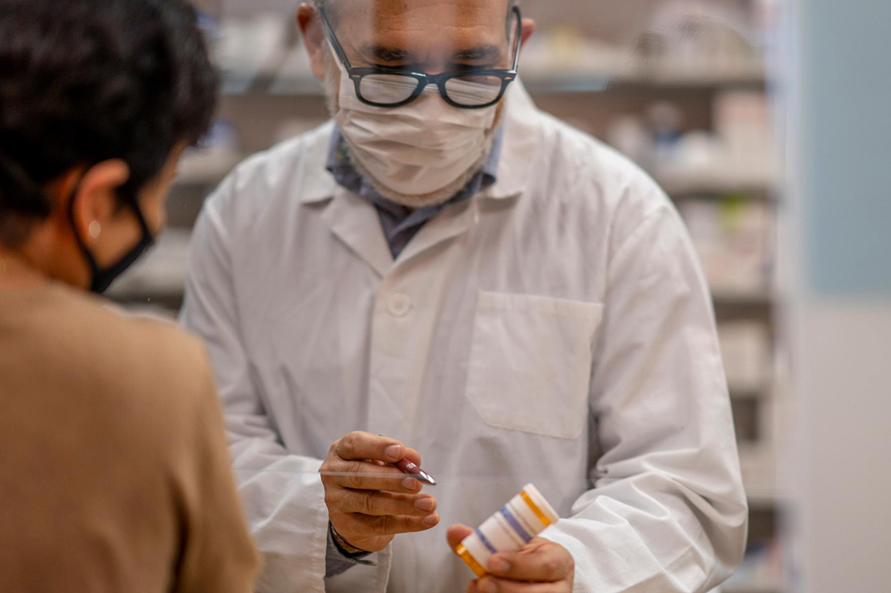 A man wearing a white coat gestures toward a pill bottle label in front of another person
