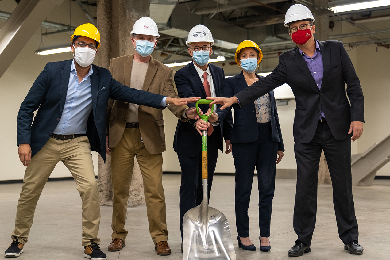 Five people wearing face masks and hard hats touch a shovel