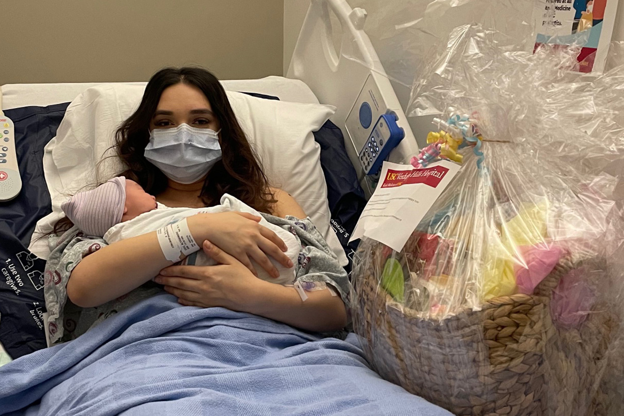 A masked woman in a hospital bed holds a baby with a gift basket next to her