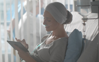 A young woman in a head scarf smiles as she uses a tablet from a hospital bed.