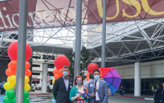 Three people stand before a Keck Medicine of USC Banner and rows of rainbow-colored columns of balloons.