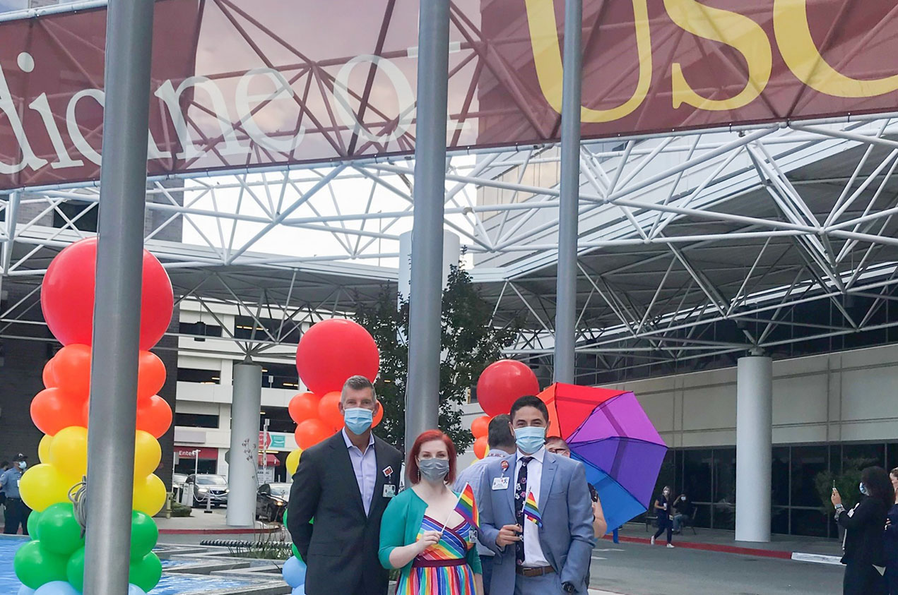 Three masked officials stand before a Keck Medicine of USC Banner and rainbow-themed columns of balloons