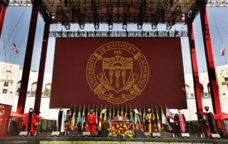 A stately commencement stage outdoors features a banner of the USC Trojan shield
