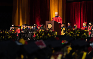 A man with bleached, spiky hair speaks at a commencement ceremony