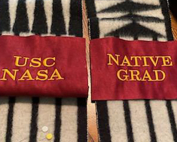 Two handwoven sashes bear satin embellishments reading, USC NASA and Native Grad