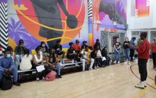 An educator addresses high school students from the side of a basketball court