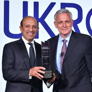A beaming doctor holds up an award beside a gray-haired man.