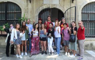 A small group of students and a professor stand in front of a centuries-old building and give the peace sign.=