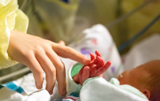 A woman's hand gently touches a premature infant's tiny, wrinkled hand.