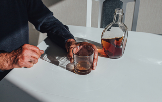 An older person's hands rest on a table, a glass of brown liquor in one hand.