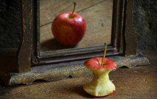 An apple core faces a mirror with a full, round apple on the other side.