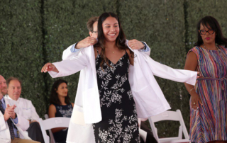 At an outdoor ceremony, a doctor puts a white coat on a beaming young woman