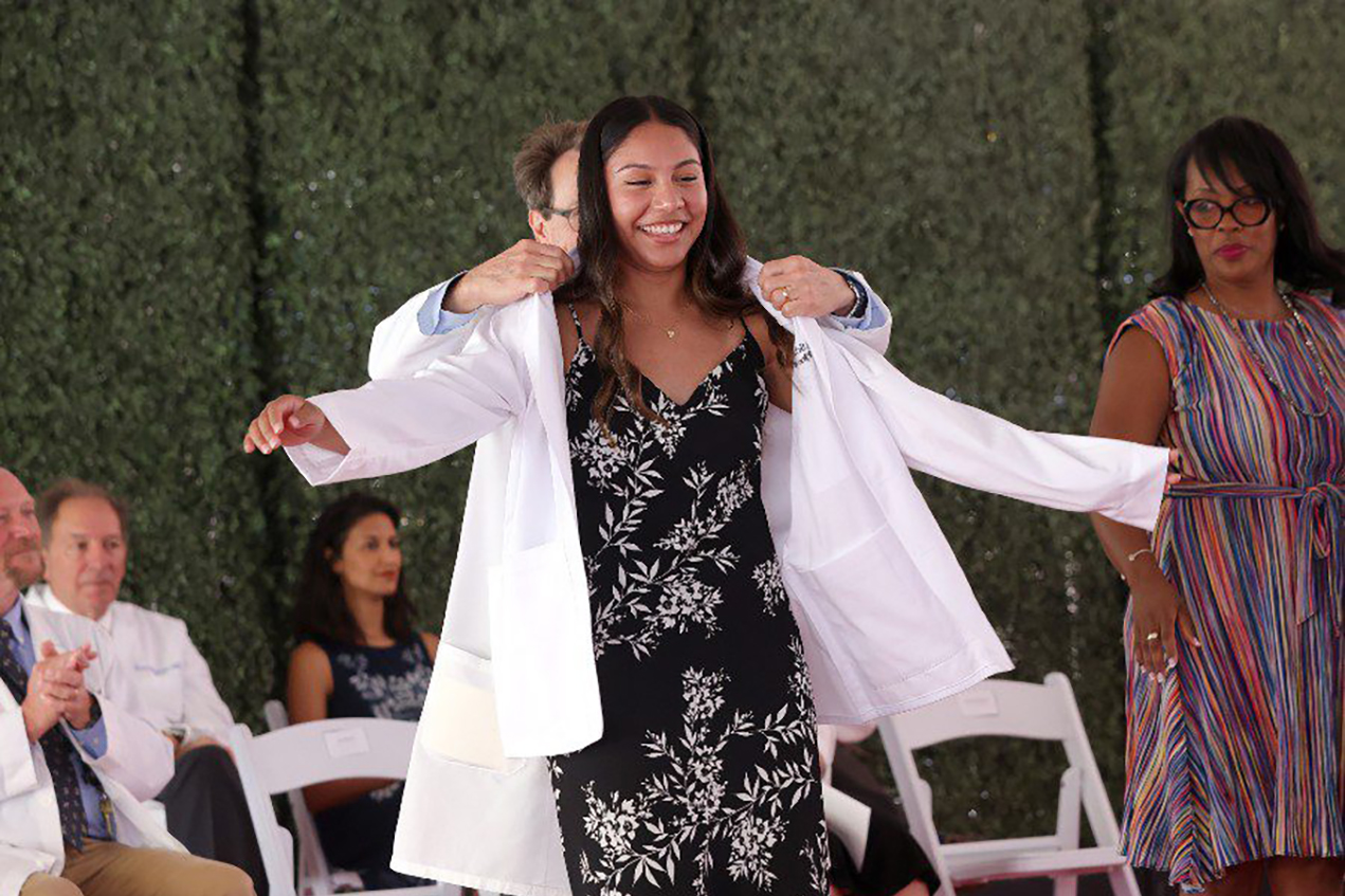 A young woman beams as a doctor fits her with a white coat