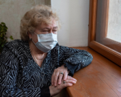 A masked older woman sits alone at a window
