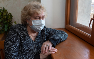 A masked older woman sits alone at a window