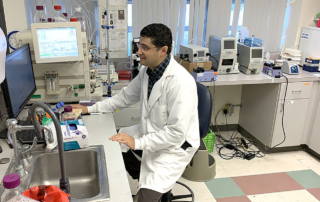 In a research lab, a bespectacled scientist in a white coat works at a computer