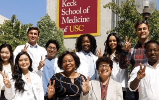Two women and a group of students hold up their hands in peace signs before a Keck School of Medicine of USC banner