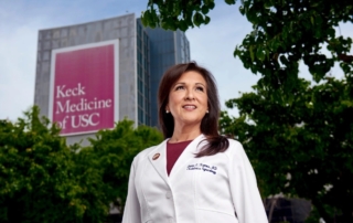 A doctor smiles proudly outside a tall building bedecked with a Keck Medicine of USC banner