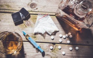 Brown liquor, pills, powder, a lighter and a syringe rest on a wooden surface.
